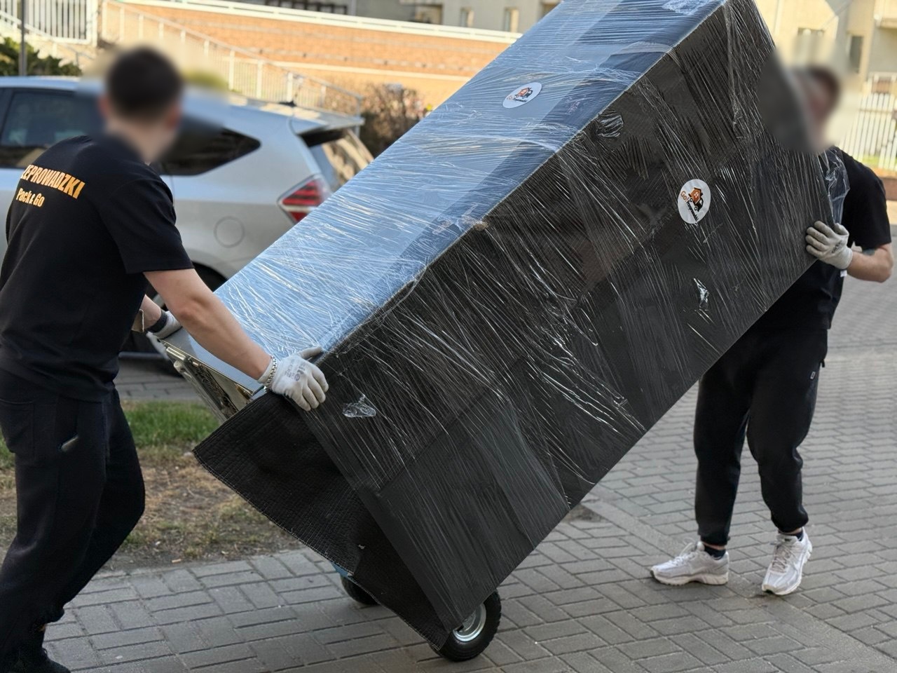 Dwóch pracowników Go Przeprowadzki transportuje zabezpieczoną folią lodówkę – bezpieczny przewóz AGD w Warszawie.