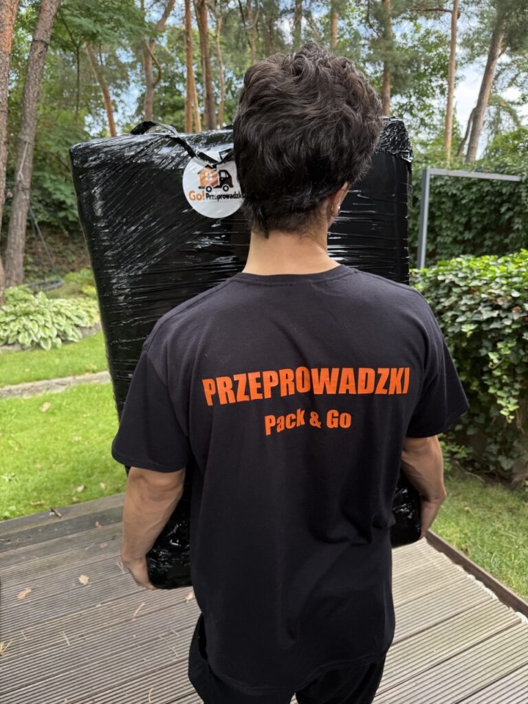 Pracownik Go Przeprowadzki w koszulce przenosi mebel zabezpieczony folią stretch z logo firmy.